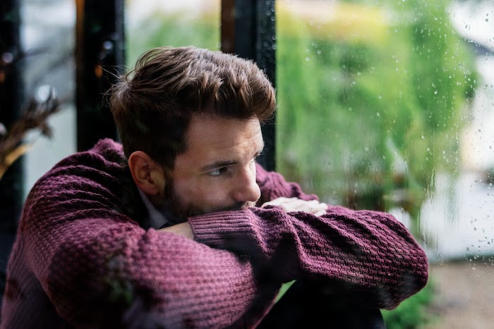 A depressed brown haired man staring out the window while it is raining